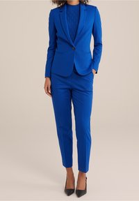 Completo su misura blu royal con giacca aderente e pantaloni affusolati. La giacca presenta un bottone singolo e una texture di tessuto liscia.