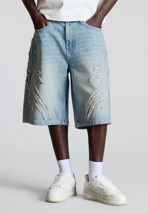 Osoba nosi svetlomodré rifľové kraťasy s textúrovanými plamennými vzormi, biele ponožky a biele tenisky, ruky v vreckách.