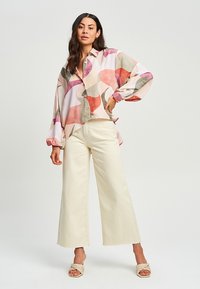 Lagani, oversized blouse s apstraktnim uzorcima u ružičastoj, crvenoj i zelenoj boji; uparen s krem hlačama širokog kroja i tkanim sandalama.