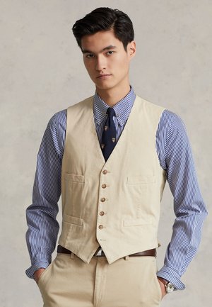 Ung mand iført beige vest, blåstribet skjorte, marineblåt mønstret slips, beige bukser og ur, stående med hænderne i lommerne.