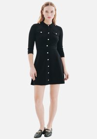 Robe en tricot noire avec un col, des manches trois-quarts, des boutons-pression et deux poches poitrine. Associée à des mocassins noirs avec une chaîne dorée.