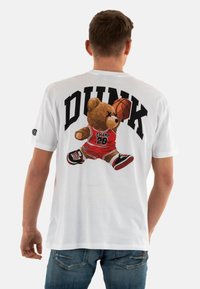Camiseta de algodón blanca con un gráfico de un oso vestido con ropa de baloncesto roja y zapatos negros, con el texto "DUNK" arriba en negro.