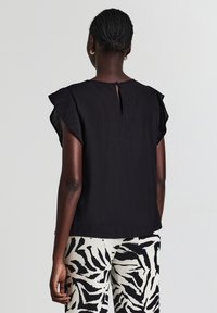 Donna con capelli intrecciati, che indossa un top senza maniche nero a ruches e pantaloni bianchi con strisce astratte nere e bianche, di spalle su sfondo neutro.