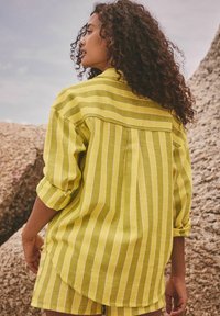 Camicia a righe gialle con maniche arrotolate e orlo leggermente curvo. Il tessuto sembra leggero con una finitura texture.