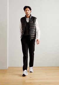 Zwarte gewatteerde vest over een grijze trui, gecombineerd met zwarte broeken en witte sportschoenen. Eenvoudig, modern ontwerp met een slanke pasvorm.