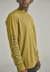 Langärmliges Shirt in Senfgelb, mit glatter Textur und Druckdesign auf dem linken Ärmel. Verfügt über einen Rundhalsausschnitt.