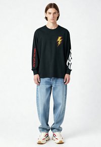 Jonge man die een zwart shirt met lange mouwen draagt met een gele bliksemschicht, rode tekst op de ene mouw en witte vlammen op de andere, lichtblauwe spijkerbroek en sneakers.