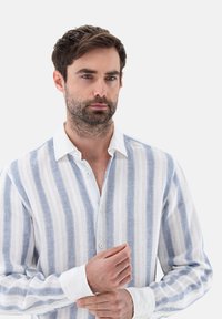 Camicia in lino con strisce verticali blu e bianche, colletto bianco e dettagli con bottoni. Maniche lunghe con polsini a contrasto.