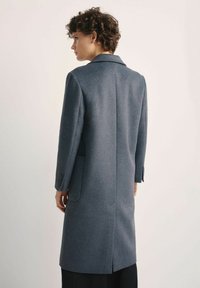 Kobieta z krótkimi kręconymi włosami w długim szarym płaszczu, stojąca tyłem z rękami swobodnie opuszczonymi wzdłuż ciała na jasnym, gładkim tle.