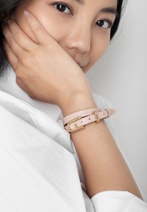 Mlada oseba nosi svetlo roza usnjen dvostranski zapestni trak z zlatim zaponko, roko ima naslonjeno na obraz, oblečena ima belo majico.