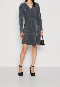 Robe portefeuille bleue à manches longues, tissu texturé, encolure en V, taille cintrée et ceinture nouée. Portée avec des chaussures à talons blocs noires.