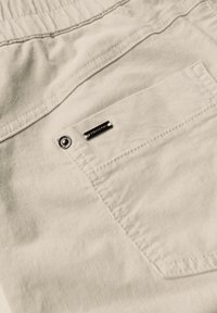 Shorts en tissu beige avec une poche arrière dotée d'un bouton argenté et d'une petite étiquette de marque rectangulaire en métal foncé.