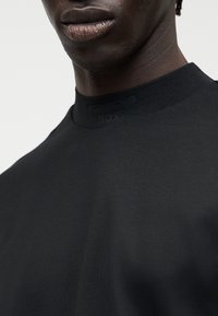 Κοντινό πλάνο ενός ατόμου που φοράει μαύρο πουκάμισο με ψηλό γιακά και διακριτικό ανάγλυφο κείμενο στον γιακά.