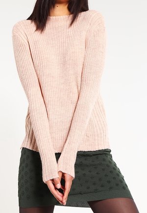 Femme portant un pull côtelé rose pâle et une mini-jupe texturée vert foncé, debout les mains jointes devant elle sur un fond blanc.