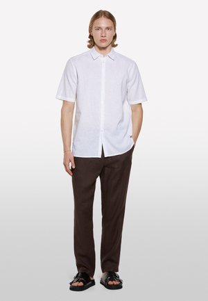 Chemise en lin blanche à manches courtes avec un col boutonné, associée à un pantalon en lin marron et des sandales noires. Design simple et décontracté.