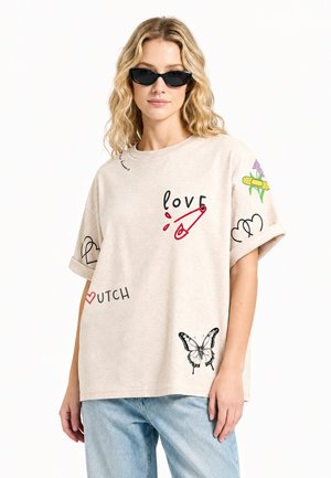Donna bionda con occhiali da sole neri e t-shirt beige oversize con grafiche di amore, farfalla, cuore e fiore, con jeans azzurri chiari.