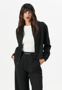 Zwarte kortjack gemaakt van gladde stof met een rits, gecombineerd met een witte t-shirt met ronde hals en zwarte wijde broek met een waistband-logo.