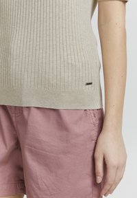 Maglione beige a costine con maniche corte e orlo dritto. Abbinato a pantaloni corti rosa, che presentano una finitura testurizzata e un piccolo etichetta accentata.