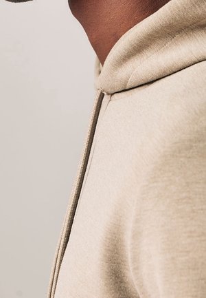 Nærbillede af en person iført en beige hættetrøje med en synlig snøre på venstre side.