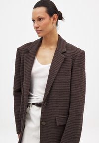 Brauner Strickblazer mit strukturierter Oberfläche, großen Revers und zwei Knöpfen. Getragen über einem weißen Oberteil und kombiniert mit einem hellen Rock.