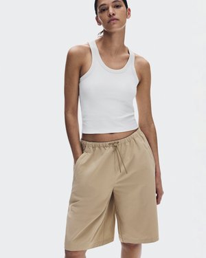 Osoba koja nosi bijelu kratku majicu bez rukava i bež kratke hlače do koljena s rukama u džepovima, stojeći pred jednoličnom svijetlom pozadinom.