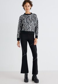Pull noir et blanc avec motif géométrique, associé à un pantalon évasé noir avec des coutures blanches. Le modèle porte des bottes à plateforme.