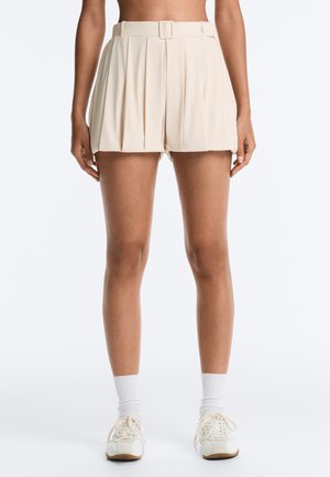 Femme portant un short plissé beige taille haute avec ceinture, des chaussettes blanches et des baskets blanches à lacets, debout devant un fond uni.