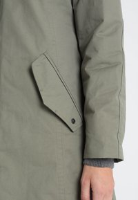 Olivově zelený kabát s elegantním designem, texturovanou látkou, kýženou boční kapsou a černým knoflíkem u vstupu do kapsy.