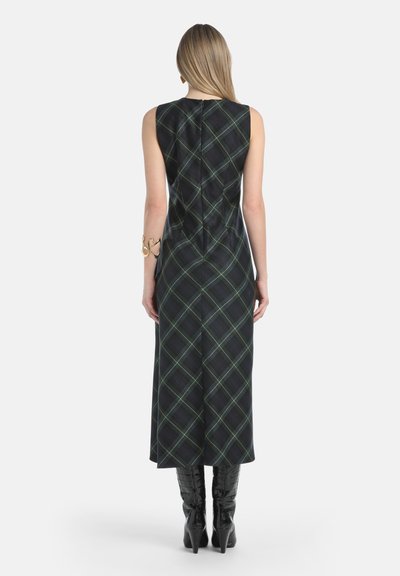 Donna che indossa un vestito midi a maniche corte in verde scuro e blu a quadri, con stivali neri fino al ginocchio, in piedi di spalle contro uno sfondo semplice.