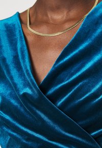 Vêtement en velours turquoise avec un décolleté en V profond et une texture douce et drapée ; accompagné d'un collier lisse de couleur or.