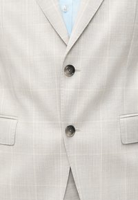 Ανοιχτό μπεζ υφασμένο σακάκι με δύο σκούρα κουμπιά, γιακά με εγκοπή και τσέπες με καπάκια, φορεμένο πάνω από ανοιχτό γαλάζιο πουκάμισο.