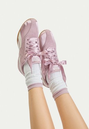 Ben med vita strumpor med rosa kant och rosa Reebok-sneakers med breda sidenband upplyfta mot en enfärgad ljus bakgrund.
