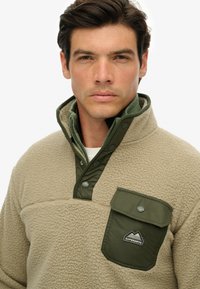 Beige fleece-pullover med grøn nylonkrave og lomme. Har trykknaplukning og fremtrædende branding på brystlommen.