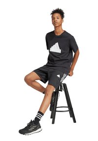Schwarzes T-Shirt mit kurzen Ärmeln und weißem Adidas-Logo, kombiniert mit schwarzen Shorts, die drei weiße Streifen haben. Schwarze Sportschuhe mit strukturiertem Design.