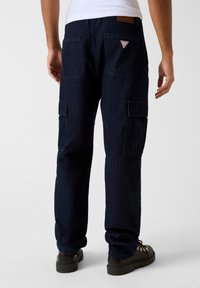 Ciemnoniebieskie spodnie cargo wykonane z denimu, z wieloma kieszeniami, skórzaną naszywką na pasku i trójkątnym logo z tyłu.