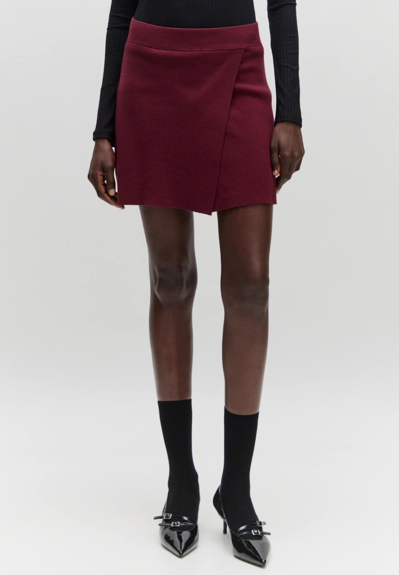 Burgunderfarbener Wickelrock mit glatter Textur und Taillenbund, kombiniert mit schwarzen knielangen Socken und glänzenden schwarzen spitzen Absätzen.
