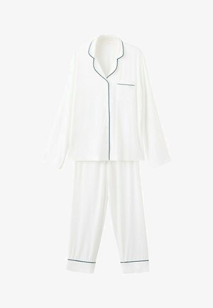 Femme aux cheveux retenus, portant un haut de pyjama blanc à manches longues avec des liserés noirs et un pantalon de pyjama blanc assorti, debout de profil.