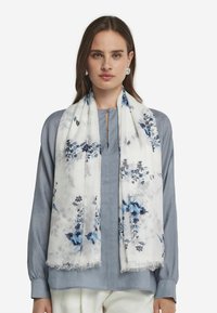 Écharpe à imprimé floral blanc, ornée de fleurs bleues et navy. Fabriquée en tissu léger avec des bords à franges, présentée sur un blouse bleu clair.