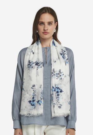 Écharpe à imprimé floral blanc, ornée de fleurs bleues et navy. Fabriquée en tissu léger avec des bords à franges, présentée sur un blouse bleu clair.