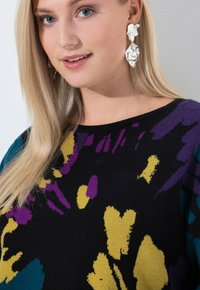 Blonde Frau mit großen silbernen abstrakten Ohrringen und einem schwarzen Pullover mit abstrakten Mustern in Gelb, Lila und Türkis.