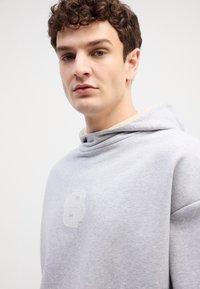 Šedá mikina s kapucí z jemného materiálu; má decentní embosované logo na hrudi a uvolněný design výstřihu.
