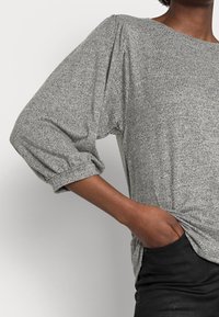 Grau, strukturiertes Langarmshirt mit Puffärmeln, lockerer Passform und rundem Ausschnitt, kombiniert mit schwarzen, figurbetonten Jeans.
