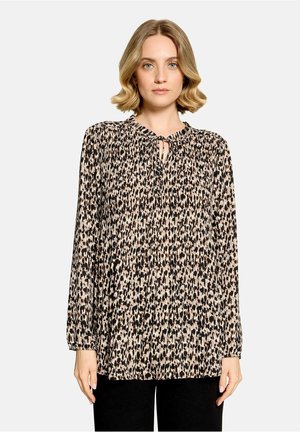 Frau mit kurzen blonden Haaren, die eine langärmelige Bluse mit Leopardenmuster und kleinem Schlüsselloch-Ausschnitt mit Schnürung trägt.