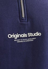 Sudadera con cremallera en color navy, fabricada con tejido texturizado, que presenta una cremallera dorada vertical y un logo en blanco que dice "Originals Studio" con texto adicional debajo.