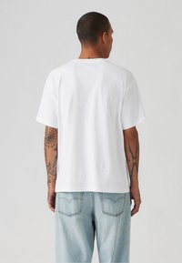 Weißes Baumwoll-T-Shirt mit lockerem Schnitt und kurzen Ärmeln, mit einem schlichten Rücken. Kombiniert mit hellblauen Jeans.