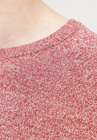 Röd och vit melerad stickad tröja med ribbad rundhalsning och texturerad tyg. Har en avslappnad design med subtila, oregelbundna mönster.