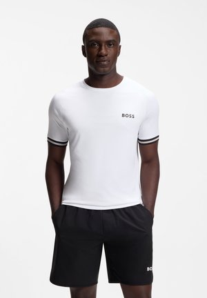 Mann trägt ein weißes BOSS-T-Shirt mit schwarzen gestreiften Ärmeln und schwarze BOSS-Shorts, steht mit den Händen in den Taschen vor weißem Hintergrund.
