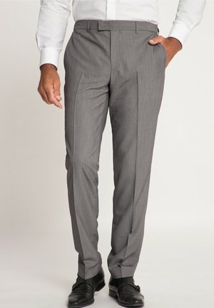 Homme portant un pantalon gris sur mesure, des chaussures formelles noires et une chemise blanche, debout avec une main dans la poche.