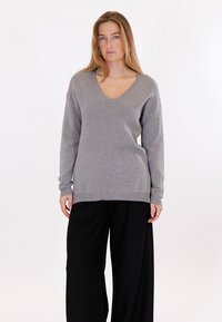 Žena s dlouhými blond vlasy, oblečená v šedém svetru s výstřihem do V a černých širokých kalhotách, stojící před jednoduchým bílým pozadím.