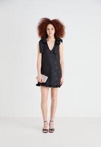 Black dress featuring a V-neck, ruffled hem, and oversized bow straps. Smooth textured fabric with subtle floral patterns. Silver clutch.